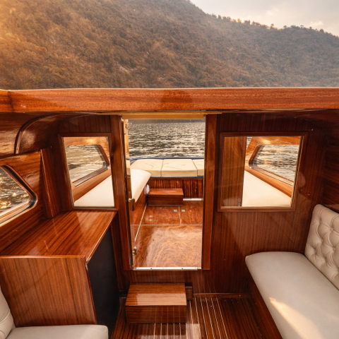 Interior of a wooden boat with beige seating, on a sunlit lake with mountains in the background.