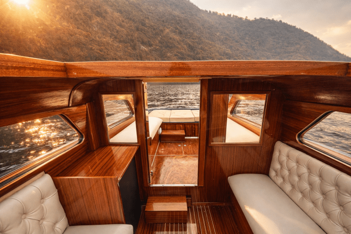 Interior of a wooden boat with beige seating, on a sunlit lake with mountains in the background.
