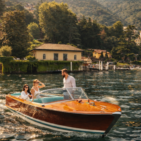 Three people on a wooden boat on a lake with scenic buildings and hills in the background.
