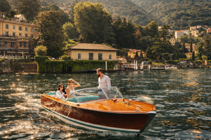 Three people on a wooden boat on a lake with scenic buildings and hills in the background.