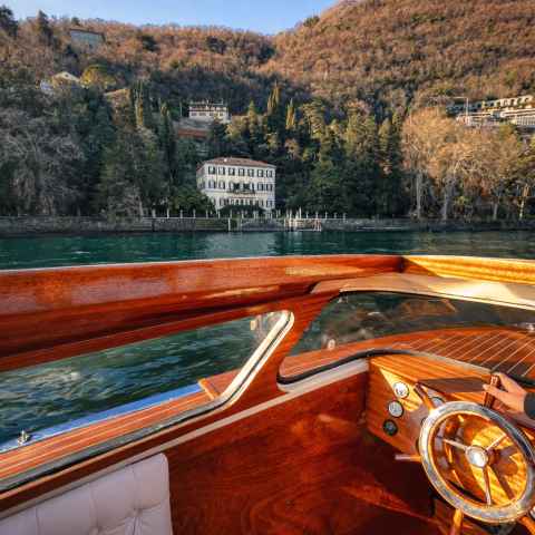 Wooden boat interior with steering wheel facing a lakeside building and forested hill.