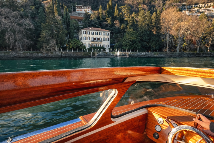 Wooden boat interior with steering wheel facing a lakeside building and forested hill.
