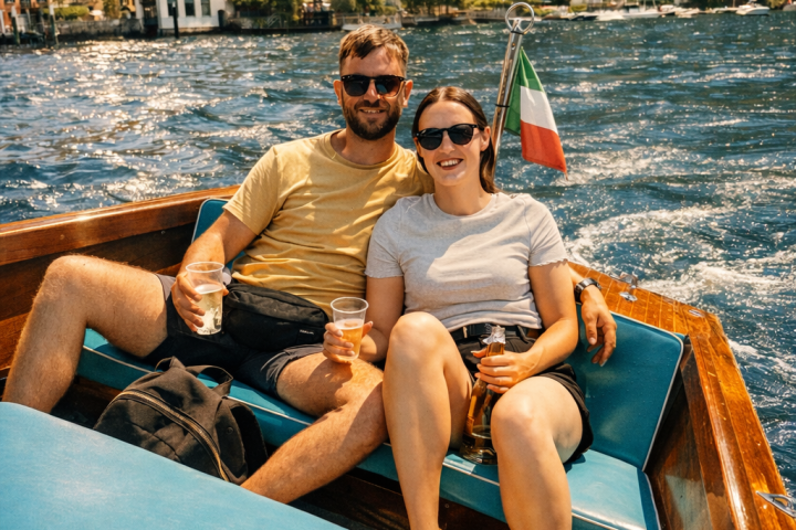 Couple relaxing on a boat with drinks, scenic hillside village in background.