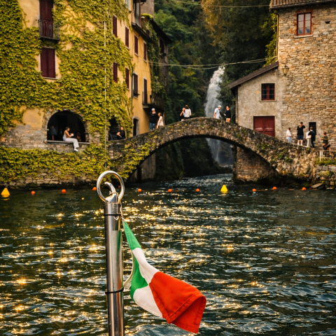 Italian flag on boat, lake view with buildings, bridge, and waterfall in background.