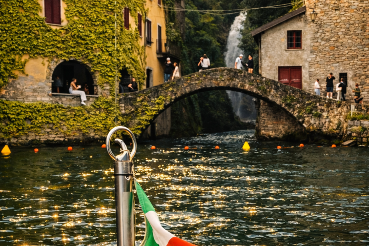 Italian flag on boat, lake view with buildings, bridge, and waterfall in background.